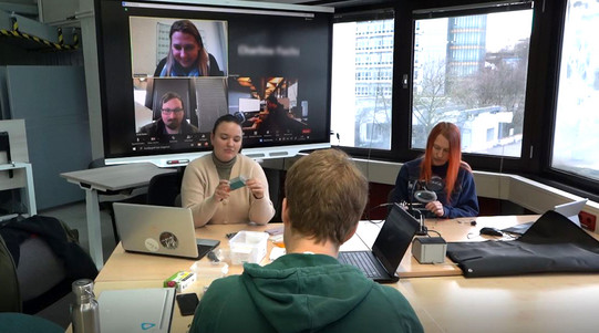 Hybride Lernsituation. Zoomkonferenz über Smartboard, davor 3 Personen arbeitend an einer Tischgruppe mit Elektrowerkzeug