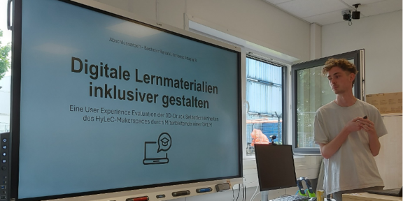 Foto des Vortrages. Man sieht den Vortragenden neben einem interaktiven Display mit seinen Präsentationsfolien. Die Überschrift des Foliensatzes lautet: "Digitale Lernmaterialien inklusiver gestalten"