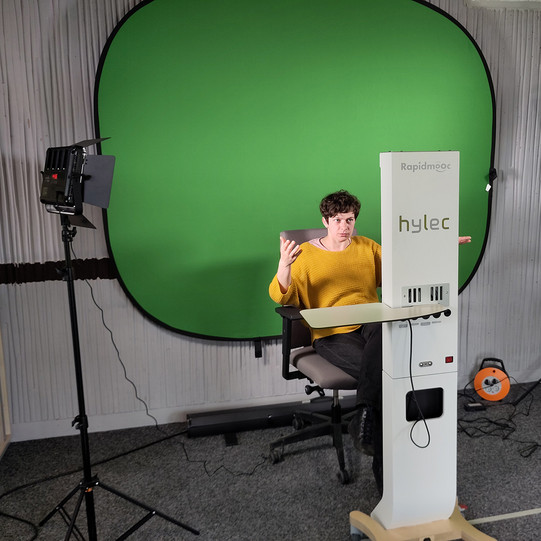 Hintergrund Greenscreen, vorne Rapidmooc, dazwischen sitzt eine Frau und benutzt die Videostation