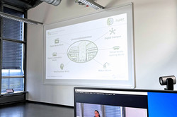 Ein Bildschirm im Vordergrund, dort drauf zu sehen eine weiblichen Person, die in einer hybriden Session etwas über das Hybrid Learning Center erzählt. Im Hintergrund eine Präsentation über das HyLeC mit einem Beamer an die Wand geworfen.