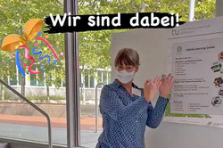 Im Vordergrund ein Infostand der Universitätsbibliothek und des Hybrid Learning Centers, weiter hinten im Bild eine weibliche Person, die vor einem Infoplakat steht und etwas erklärt. 