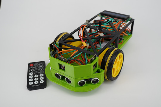 Side standing model car. Light green front with four round sensors on the bottom. Two black and yellow tires on the sides of the car. Dense wiring inside the car visible from above. Black remote control with round white buttons to the left of the car.