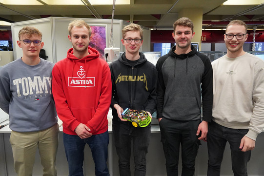 Facing the camera, the five students stand side by side in front of the 3D printers. The student in the middle is holding the model car in his hand.
