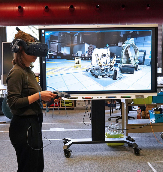Smartboard im Raum E1 mit einer jungen Frau davor, die eine virtual Reality Brille trägt und spielt. Auf dem Bildschirm wird wiedergegeben, was die Frau mit der Brille sieht.