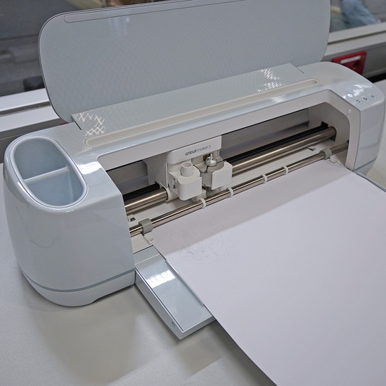 Cricut auf einem Tisch, aufgeklappt und in Aktion mit einem in Klebefolie geplottetem HyLeC-Logo. 
