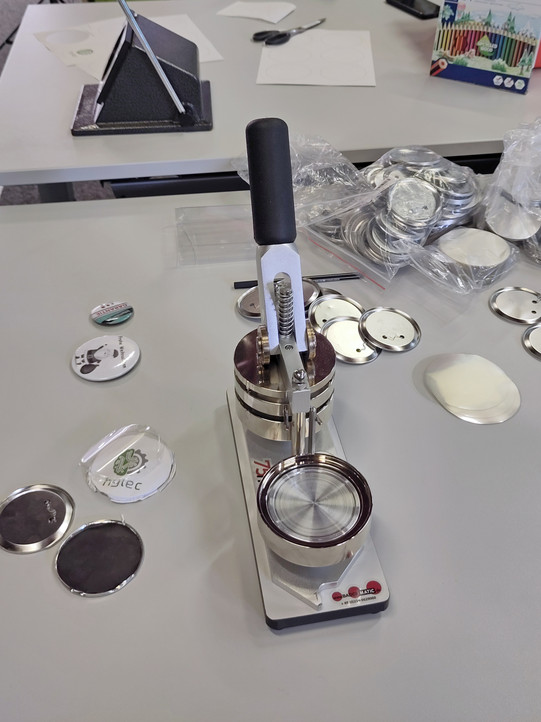 Buttonmaschine mit Zubehör auf einem Tisch. Papierstanze im Hintergrund.