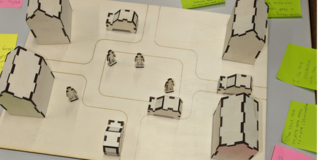 Holzplatte mit eingezeichneter Straße, darauf bewegliche 3D Holzhäuser, Autos und Männchen. Alles aus dem Lasercutter gebastelt. Rundherum liegen Post-Its mit Notizen