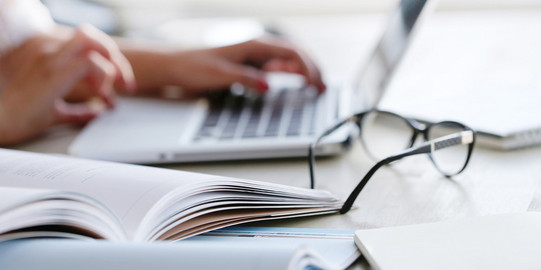 Detailfoto eines Schreibtisches. Darauf liegen Hefte, eine Brille und ein Laptop. Man sieht die Hände einer Frau, die an dem Laptop auf der Tastatur tippen.