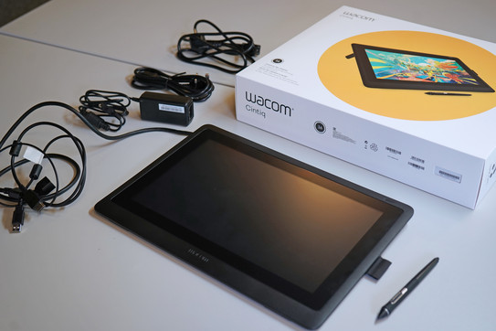 Wacom Tablet unten im Bild, darüber die Verpackung mit Beispielbild zur Nutzung und links daneben verschiedenes Zubehör, Kabel, etc.