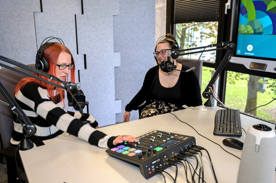 Zwei junge Frauen mit Kopfhörern vor professionellen Mikrofonen nehmen einen Podcast auf