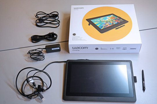 Wacom Tablet unten im Bild, darüber die Verpackung mit Beispielbild zur Nutzung und links daneben verschiedenes Zubehör, Kabel, etc.